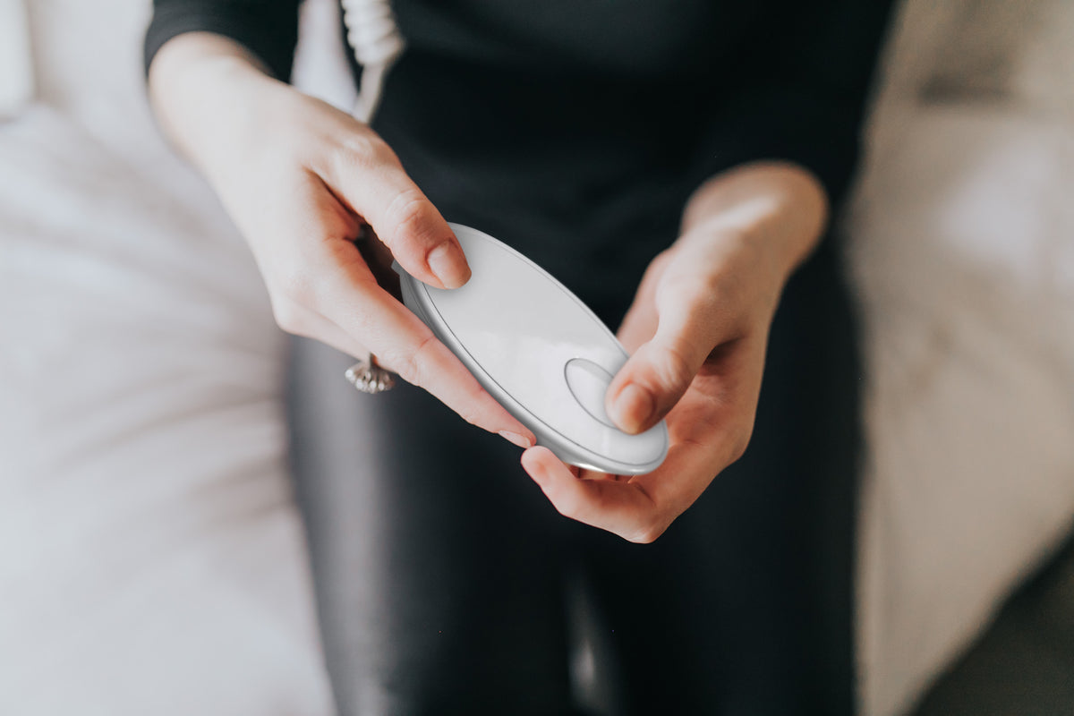 A person in black clothing holds the Rio faceLITE beauty boosting LED face mask, a white oval device with a single button, while sitting on a bed—designed to energize and rejuvenate ageing skin.