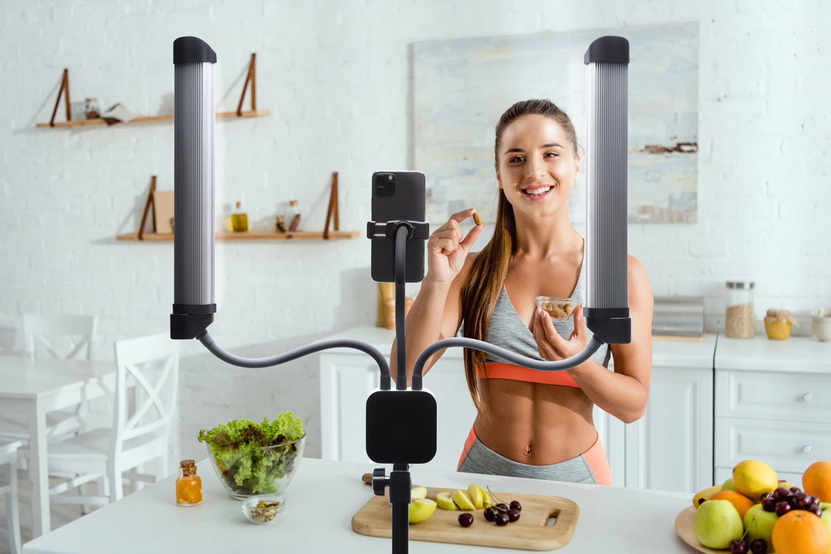 A woman in athletic wear smiles in a bright kitchen, recording herself with her smartphone while using Rio’s Professional Twin LED Strip Lights—now on sale—for high-quality content creation. Fresh fruits and vegetables are on the counter.