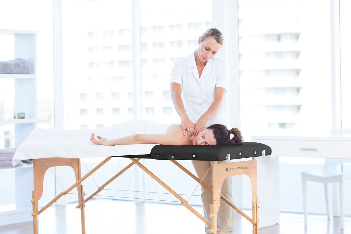 A woman enjoys a massage from a therapist in a bright, modern room, lying face down on the Rio Professional Ultra-Light Portable Massage Table covered with crisp white linens.