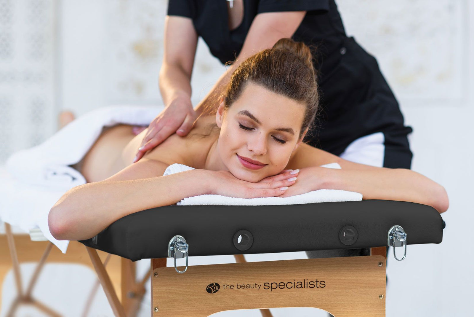 A woman enjoys a back massage on the Rio Professional Ultra-Light Portable Massage Table, smiling with her eyes closed. The table displays the Rio logo on its side, emphasizing its use for expert massage treatments.
