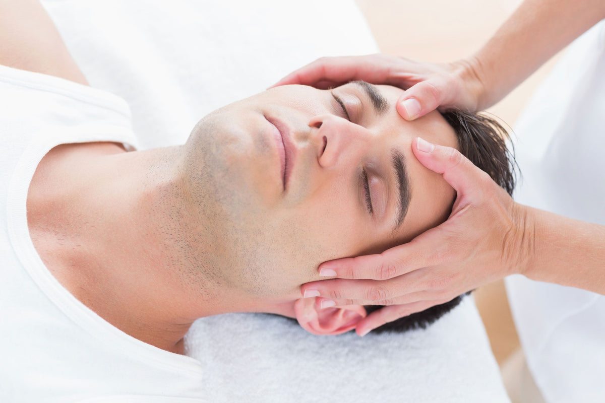 A man with dark hair, wearing a white tank top, relaxes with his eyes closed on a Rio Professional Ultra-Light Portable Massage Table as hands gently massage his temples and forehead.