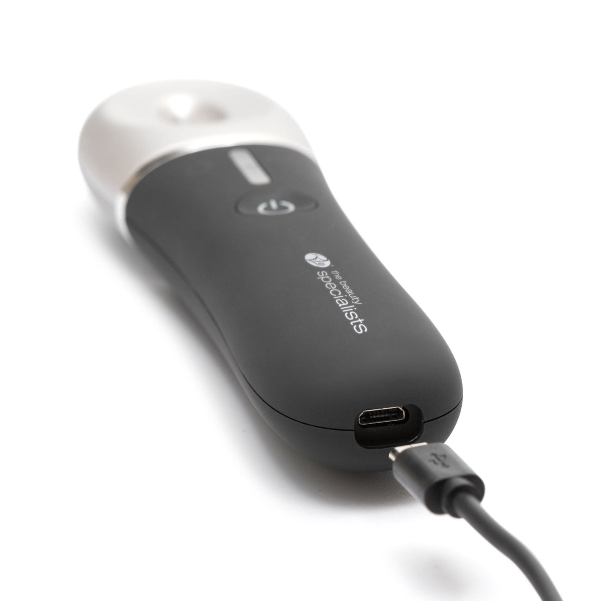 A close-up of the black Revive & Refresh device by Rio with a white top, showing its power button and USB cable attached for charging. This sleek skin-refreshing device rests elegantly on a white surface.