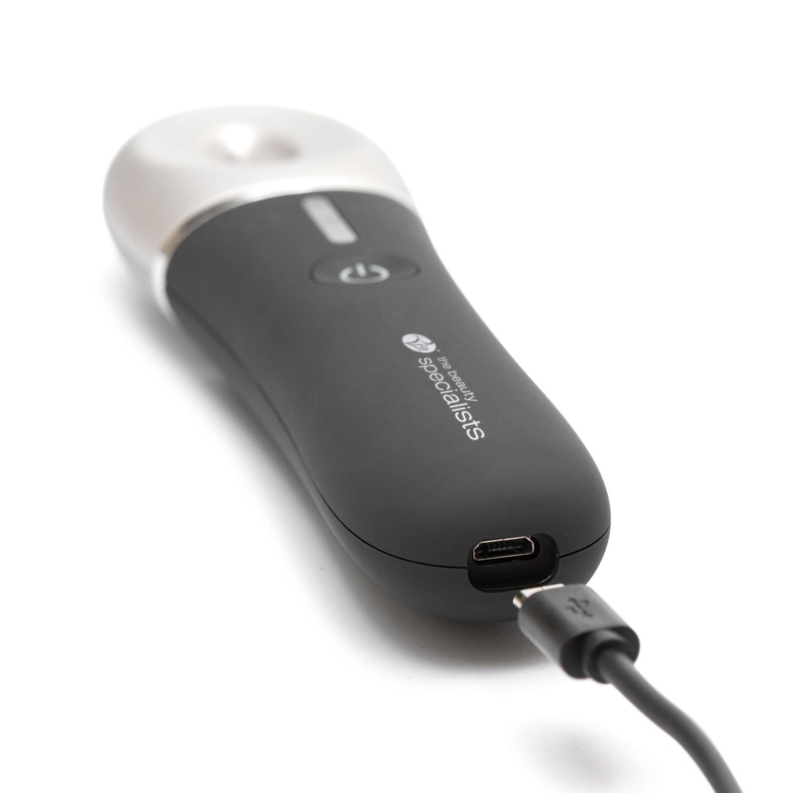 A close-up of the black Revive & Refresh device by Rio with a white top, showing its power button and USB cable attached for charging. This sleek skin-refreshing device rests elegantly on a white surface.