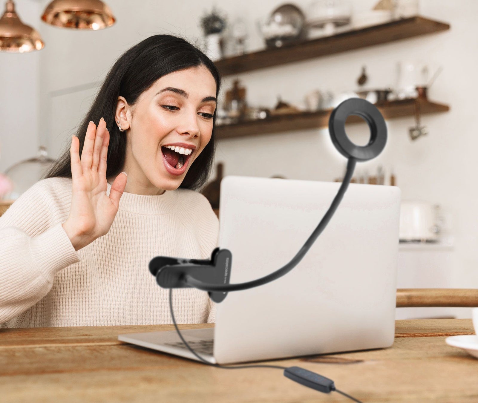A woman video calls from her cozy kitchen, smiling and waving at her laptop. The Rio Beauty Station LED Ring Light, attached to the screen, provides flattering illumination for the webcam.