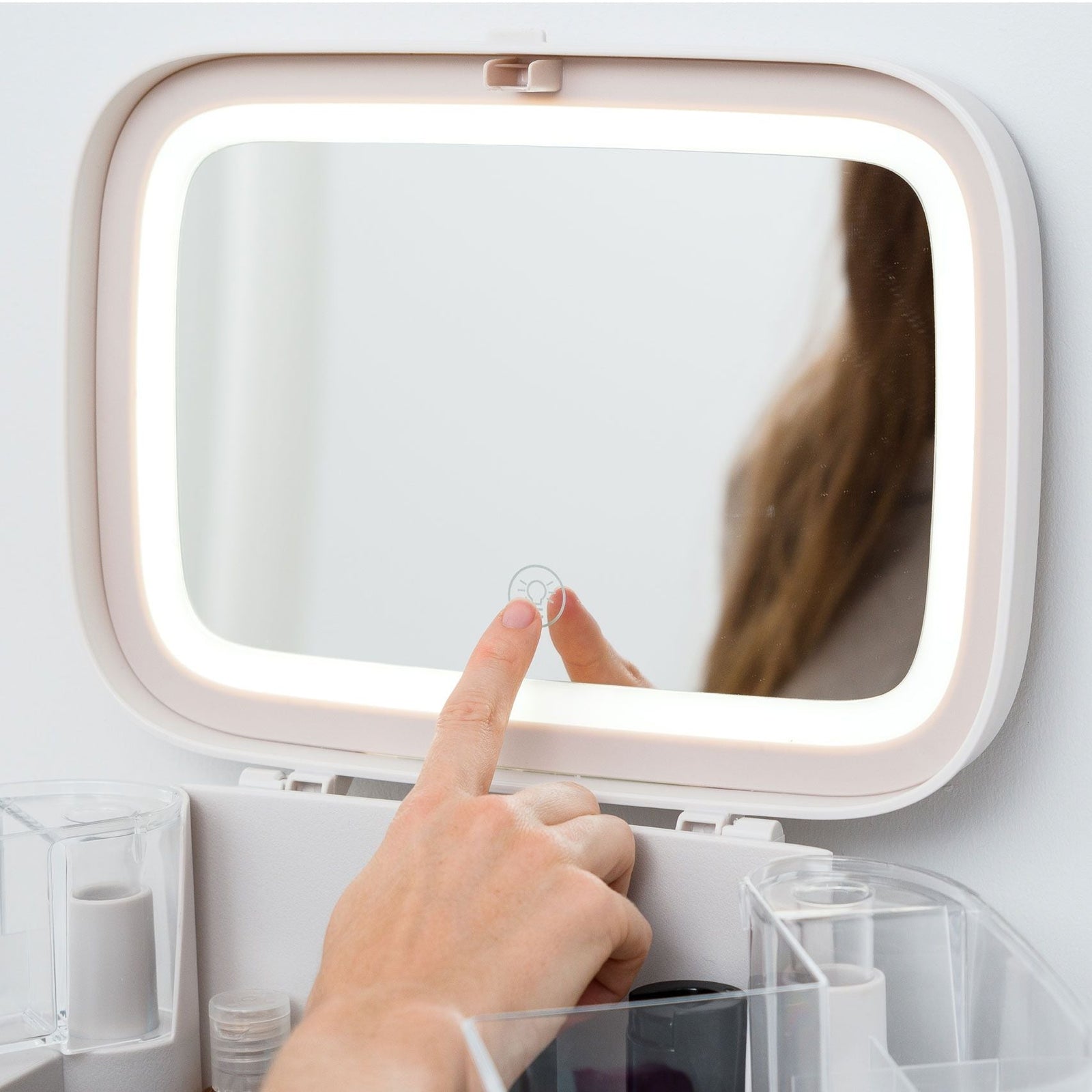 A hand touches the lit on/off button of the Rio Ultimate Beauty Storage Box With Dimmable Mirror, with a makeup organizer and containers below, and a partial reflection of a person with long hair visible.