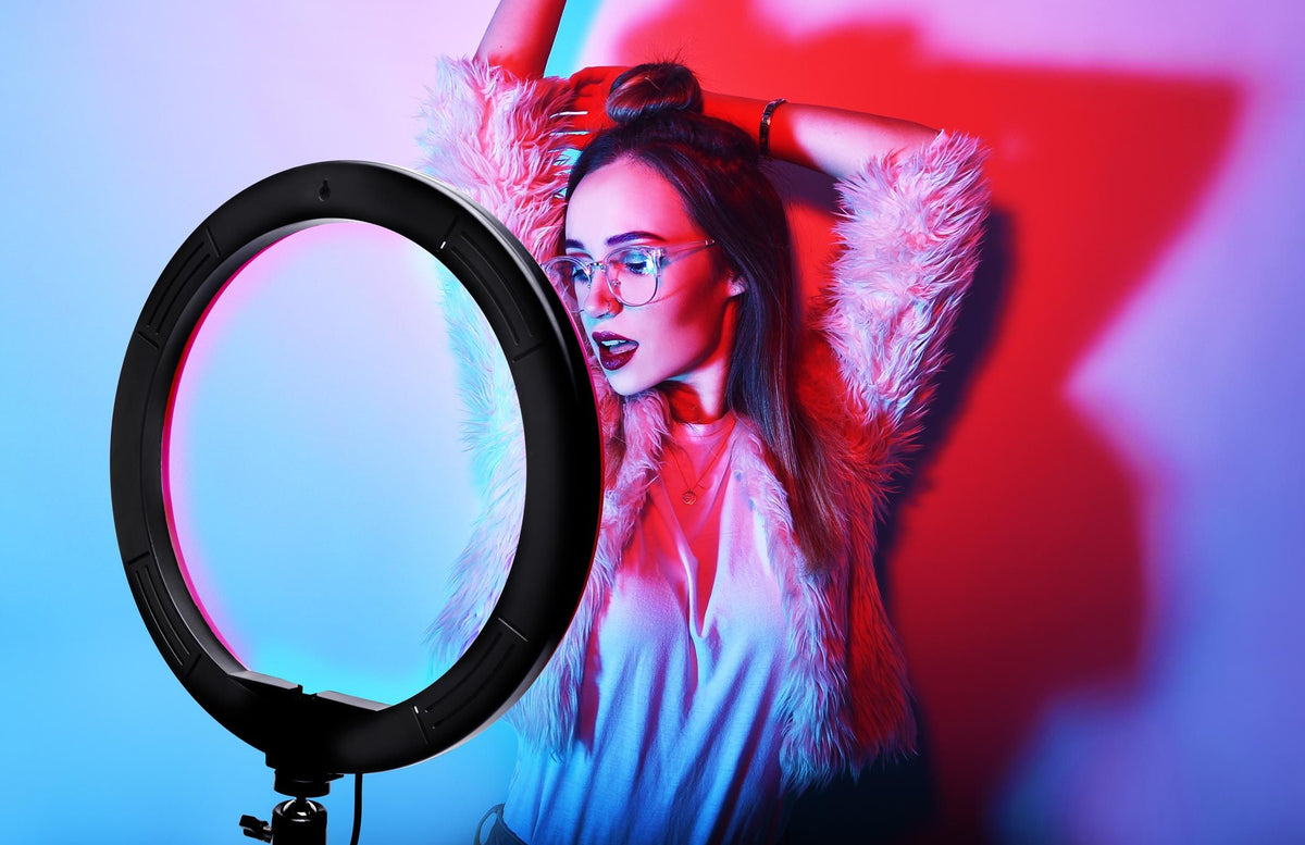 A stylish woman poses with arms up in front of the Rio Studio RGB Beauty Ring Light, wearing glasses, a furry jacket, and a white shirt. Vibrant blue and pink lighting highlights the flair of colour photography.