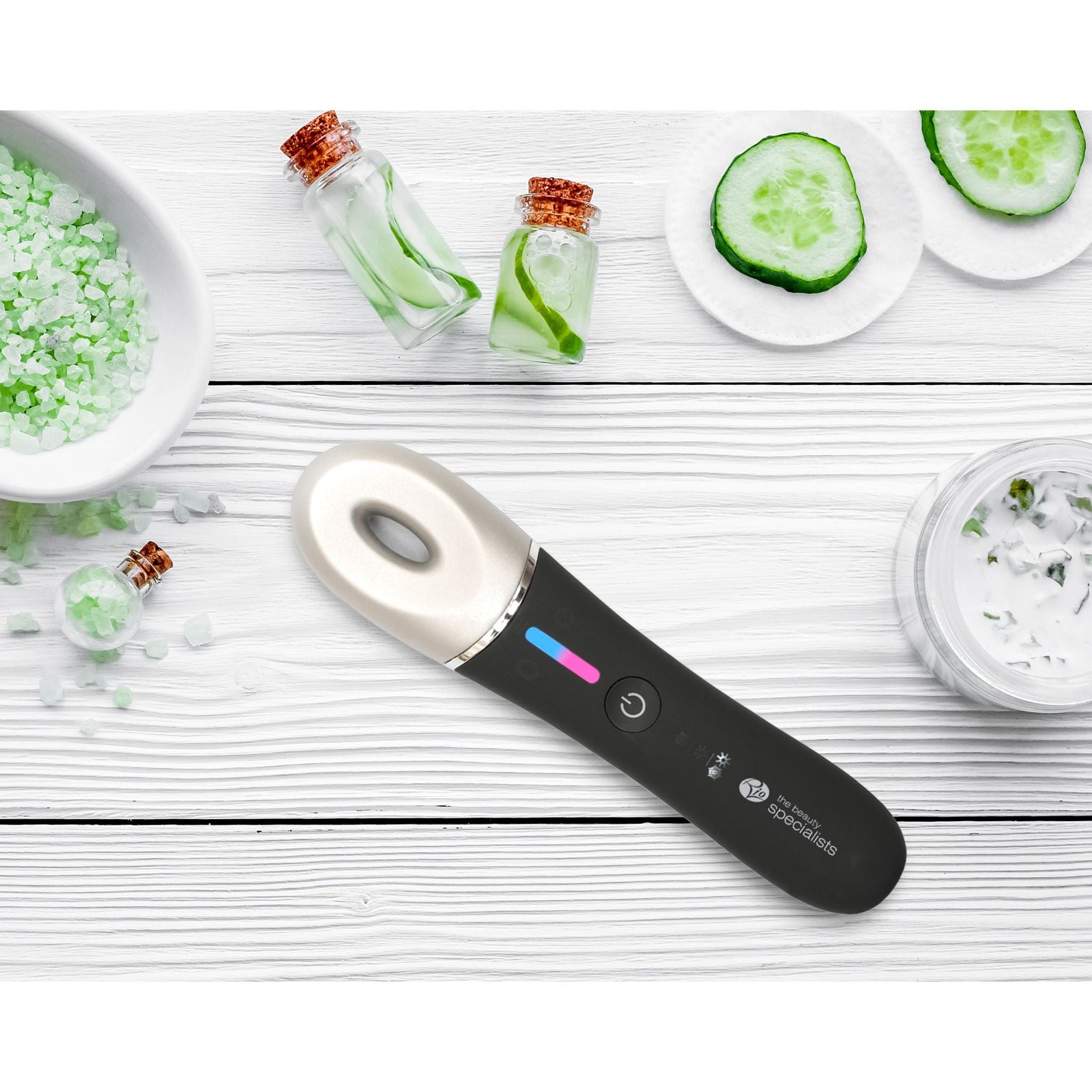 The Rio Revive & Refresh, a black and silver facial massage device, rests on white wood with green bath salts and cucumber slices—ready to soothe your skin with a creamy skincare product containing cucumber pieces.
