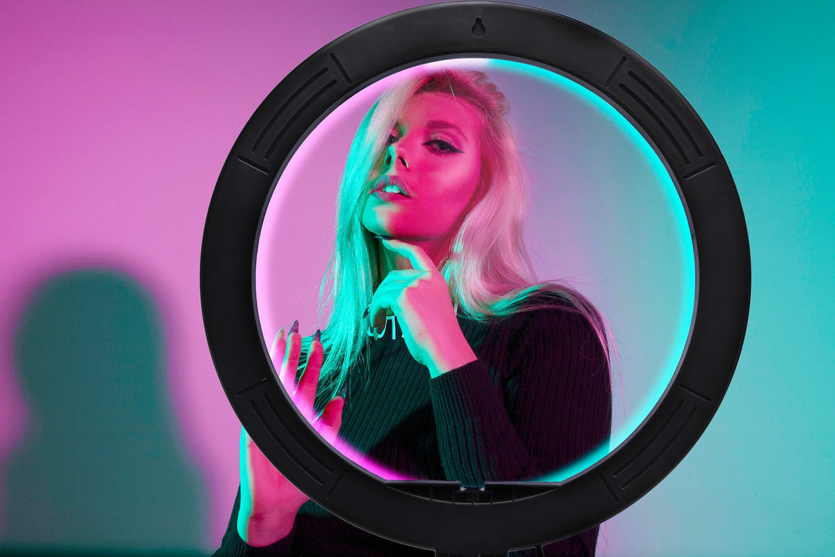A woman with long blonde hair poses confidently behind the Rio Studio RGB Beauty Ring Light, as pink and teal lighting cast vibrant shadows on her face and the wall. She touches her chin and looks at the camera in this striking, colorful scene.