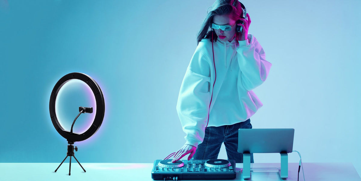 A DJ wearing headphones and sunglasses mixes music on a controller, standing at a table with her laptop and the Rio Studio RGB Beauty Ring Light, which enhances the vibrant blue and purple colours of her studio.