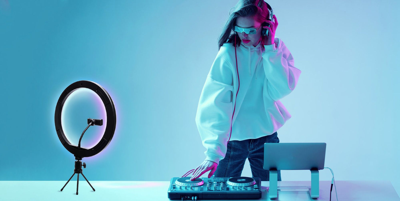 A DJ wearing headphones and sunglasses mixes music on a controller, standing at a table with her laptop and the Rio Studio RGB Beauty Ring Light, which enhances the vibrant blue and purple colours of her studio.