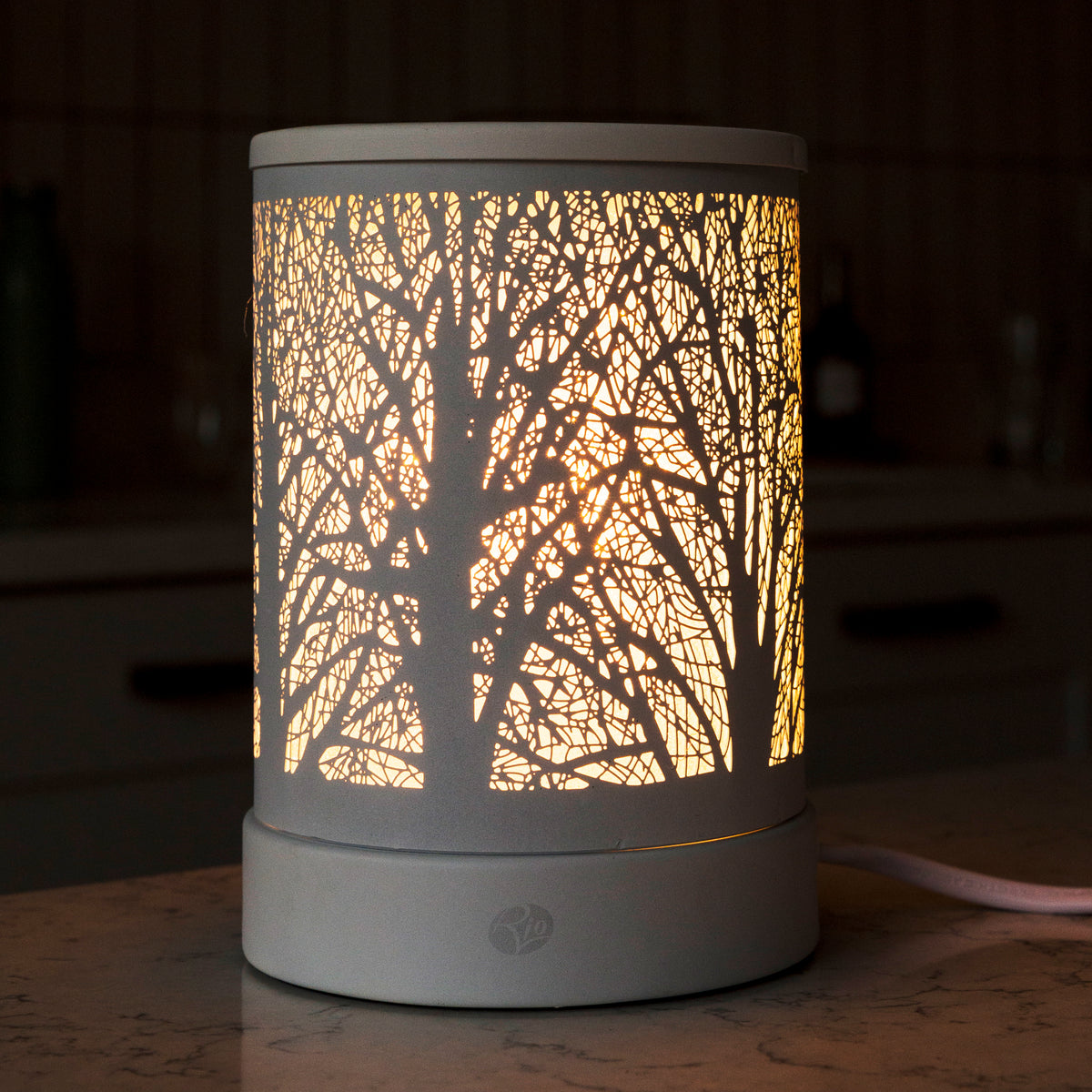 The Rio FORA Wax Melt Burner & Aroma Diffuser Lamp features a cylindrical shade with an intricate tree branch pattern, emitting a warm glow—ideal for creating a stylish, relaxing ambiance on any surface.