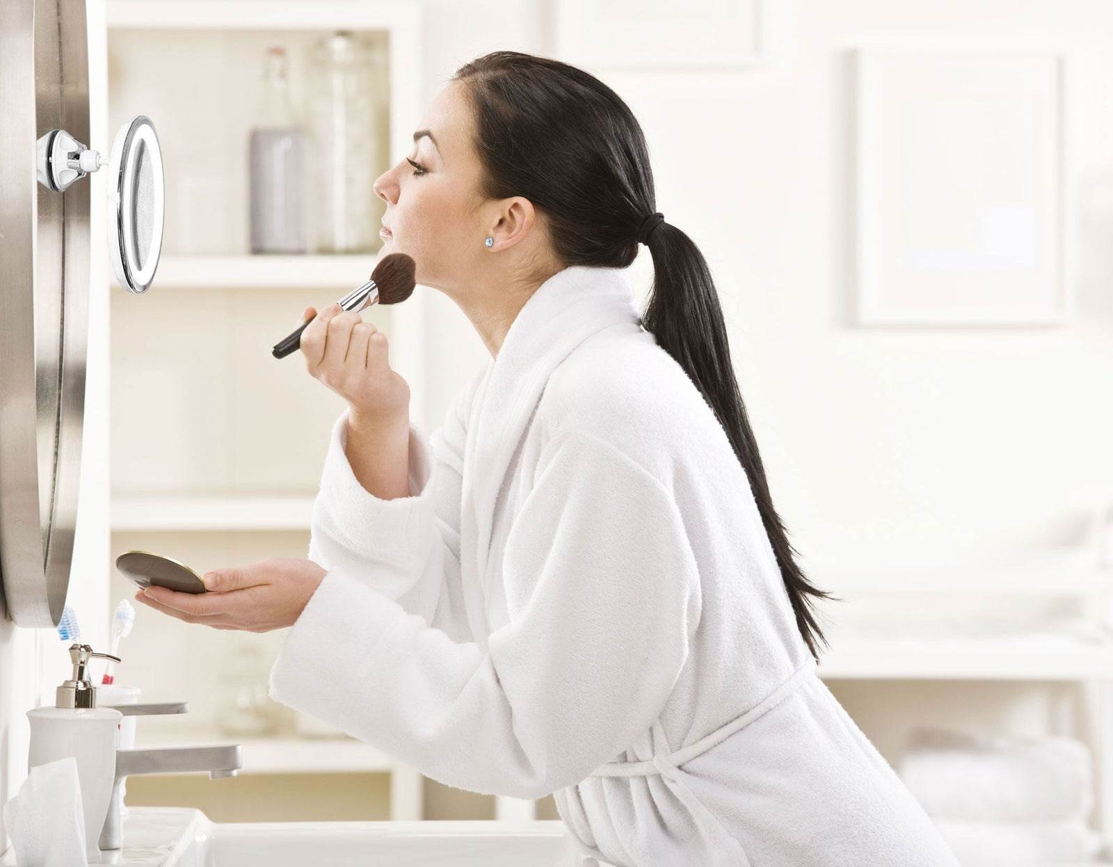 lady using Multi use LED illuminated make up mirror with suction cup base attached to larger wall mounted mirror in bathroom applying make up 