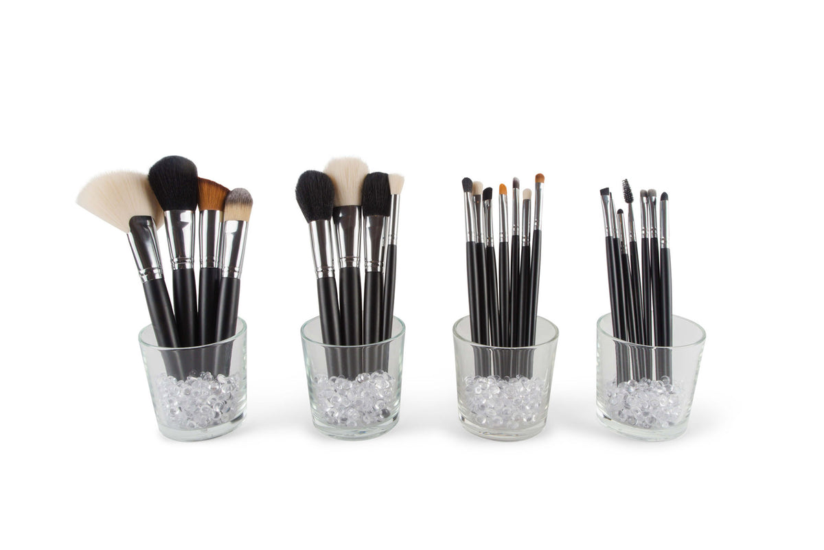 Four glass cups filled with clear beads, each displaying the Rio PROFESSIONAL SALON MAKEUP BRUSH COLLECTION & BELT—black-handled brushes of various shapes and sizes—are arranged in a row on a white background.